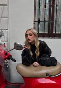 Czerwony skuter z przezroczystą szybą, beżowym siedzeniem i chromowanym lusterkiem. Czarna torebka na siedzeniu. Miękkie, falowane blond włosy, ciemne ubranie.