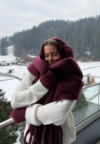 Bufanda de lana color granate envuelta alrededor de una persona que lleva un su�éter de punto color crema con puños acanalados, contra un fondo exterior nevado de árboles.