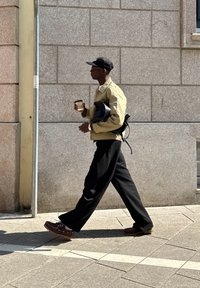 Pessoa com um casaco bege claro, calças pretas e sapatos castanhos, segurando uma caneca de café e uma bolsa preta enquanto caminha numa calçada pavimentada.