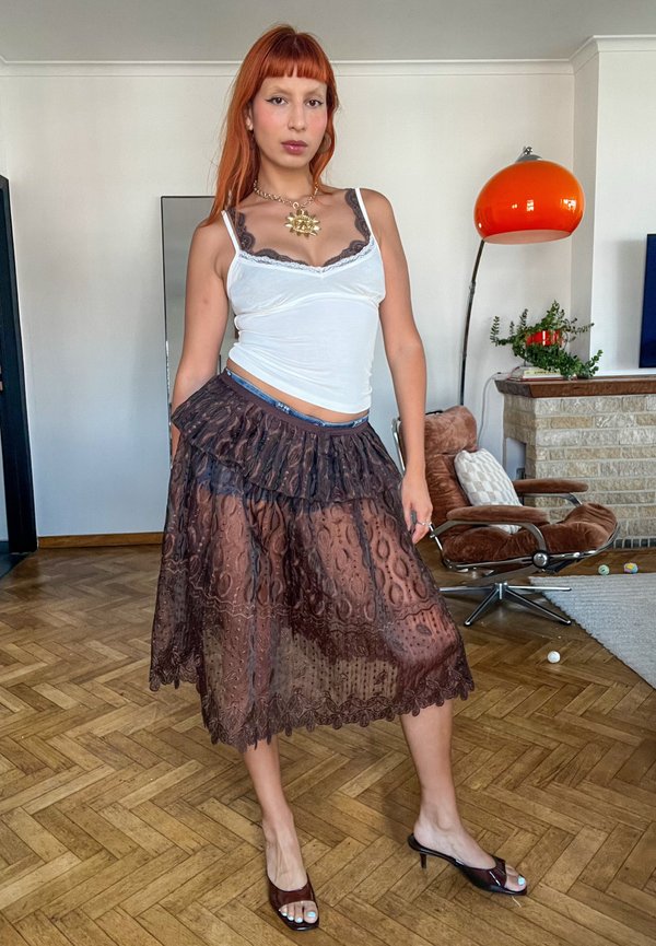 Person mit langen roten Haaren, die ein weißes Tanktop, einen mehrschichtigen schwarzen Spitzenrock und offene Pumps trägt, steht in einem Wohnzimmer mit Stuhl und Lampe.