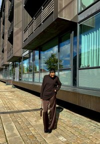 Maglione oversized marrone abbinato a pantaloni larghi in satin marrone scuro, scarpe nere e una borsa piccola. Un edificio moderno si riflette nel vetro circostante.