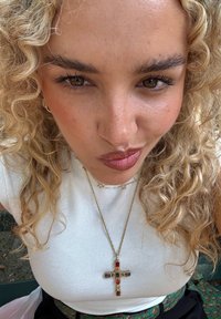 Krøllete blondt hår rammer inn et nærsyn av et ansikt som har på seg en hvit ribbet topp. Et gullkjede med et fargerikt korsanheng er sentralt.