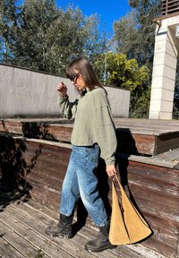 Sweater cortado verde oliva, jeans de perna larga azul clara, botas pretas e uma mala de camurça bege. Ambiente natural com um deck de madeira e árvores.