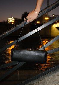 Bolso de mano de cuero negro con forma rectangular, que presenta un cierre metálico y una correa delgada, contra un fondo nocturno.