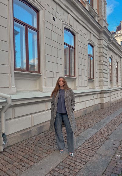 Abrigo gris texturizado sobre una camisa oscura, combinado con vaqueros de pierna ancha y tacones plateados. El fondo presenta paredes de piedra y ventanas.