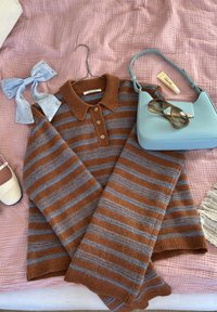 Brunstribet sweater med blå og grå mønstre, en lyseblå håndtaske, en lyseblå sløjfe, solbriller og en læbepomade på et lyserødt tekstureret tæppe.