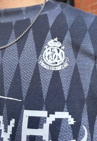 Navy blue shirt with a diamond pattern and a white embossed logo featuring a crown. Textured fabric with a smooth finish. Silver chain visible.