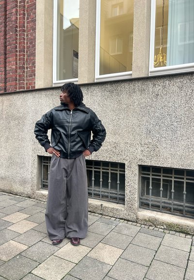 Chaqueta bomber de cuero negro con puños acanalados, llevada sobre pantalones anchos grises. De pie sobre la acera cerca de una pared texturizada y ventanas.