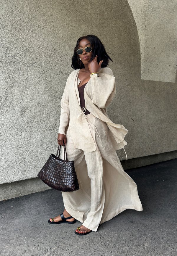 Completo in lino color beige chiaro con pantaloni larghi e un lungo giubbotto aperto. Borsa a tracolla intrecciata scura e sandali neri con dettagli dorati. Occhiali da sole indossati.