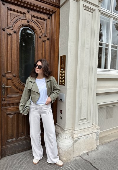 Chaqueta cropped de un verde oliva claro, camiseta blanca y pantalones de pierna ancha color crema. De pie cerca de una puerta de madera con un panel decorativo y un intercomunicador.