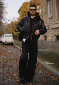 Sort læder bomberjakke med pelskrave, mørk trøje og brede sorte bukser. Model iført solbriller og skinnende sorte sko på en brostensbelagt sti.