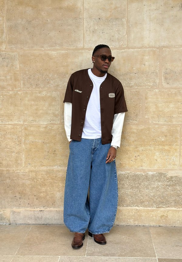 Mann trägt braunes kurzärmeliges Hemd über weißem langärmeligen Hemd, weite blaue Jeans, braune Schuhe, Sonnenbrille, lehnt an einer Steinmauer.