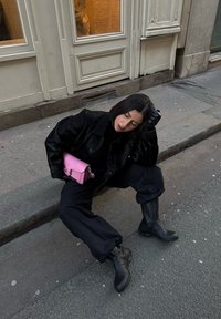 Schwarze Kunstfelljacke, schwarzer Rollkragenpullover, lockere schwarze Hose und schwarze kniehohe Stiefel. Hält eine rosa Handtasche mit silbernem Beschlag.
