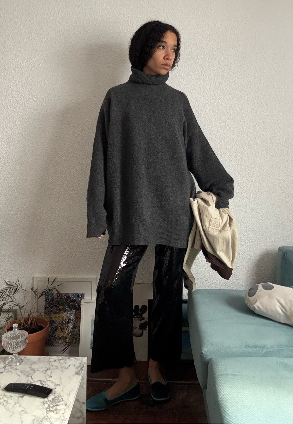 Personne debout à l'intérieur portant un pull à col roulé sombre et oversized, un pantalon noir brillant, des chaussures bleu sarcelle, tenant une veste beige à côté d'elle.