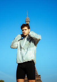 Svetlo šedý vetrovka s kapucňou a oranžovými akcentmi, nosená cez čierne šortky. Jasná obloha a Eiffelova veža v pozadí.