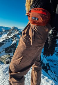 Persoon die bruine wandelbroek, zwarte handschoenen en een rode Patagonia heuptasje draagt, staand op een besneeuwde berg met rotsachtige pieken op de achtergrond.