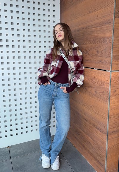 Chaqueta de forro polar a cuadros rojos y blancos, blusa burdeos, jeans azul claro, zapatillas blancas, posando frente a una pared de madera con un fondo estampado.