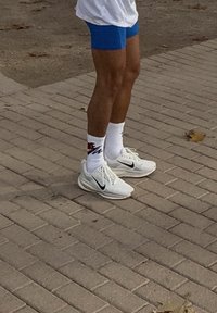 Personne portant un short bleu, des baskets Nike blanches et des chaussettes Nike blanches, debout sur une surface pavée extérieure.