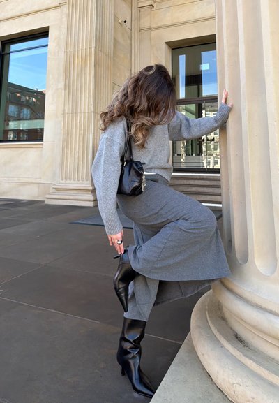 Mujer con suéter gris y falda ajusta una bota negra de tacón mientras se apoya en una gran columna beige fuera de un edificio.