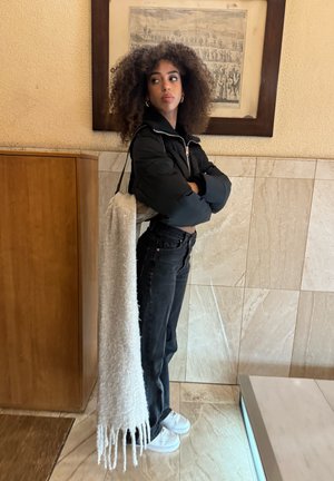 Chaqueta negra acolchada, vaqueros negros de cintura alta, zapatillas blancas y un bolso blanco texturizado con flecos. El fondo presenta azulejos beige y madera.