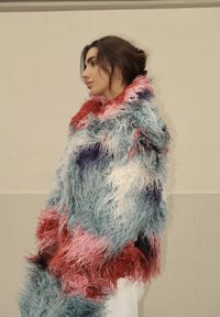 Színes, bolyhos szőrme kabát vörös, kék, rózsaszín és fehér árnyalatokkal; magas gallér; texturált, hosszú szálak; fehér ruházat fölött viselve.