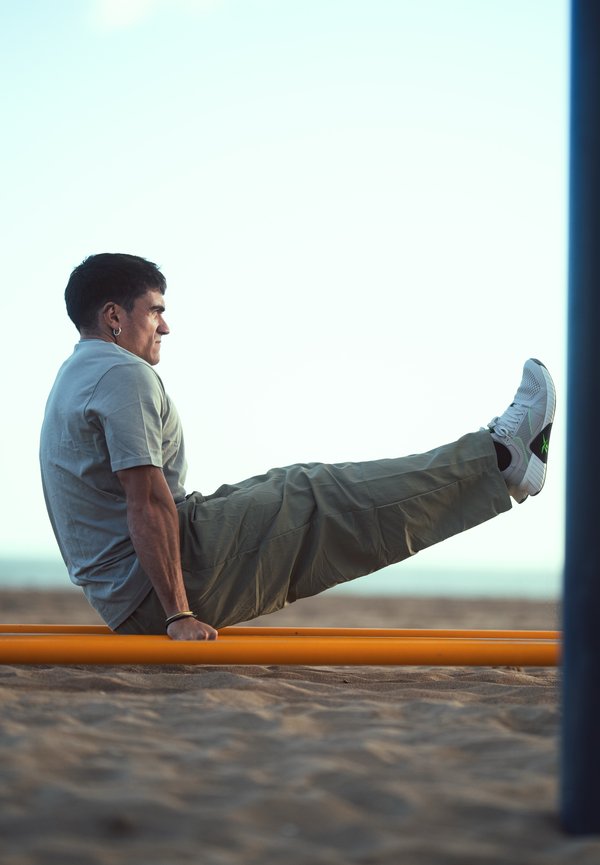 Mężczyzna w szarej koszulce i spodniach wykonuje ćwiczenie L-sit na pomarańczowych, równoległych poręczach na zewnątrz, na piaszczystym podłożu.