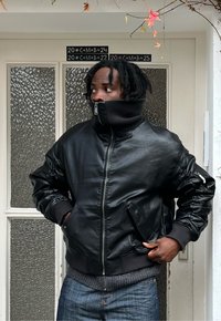 Vīrietis ar drediem, ģērbies melnā ādas jaku ar rāvējslēdzēju un zilās džinsās, stāv ar rokām kabatās un skatās pa kreisi pie teksturētas stikla durvīm.