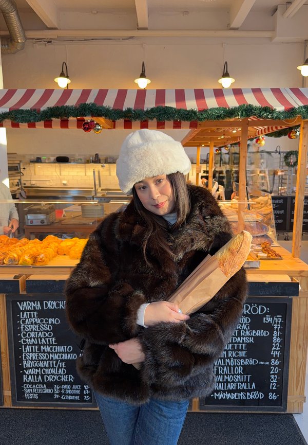 Kvinde iført pelsjakke og hvid hat, der holder en baguette i en bagerbod dekoreret med guirlander og stribet baldakin.