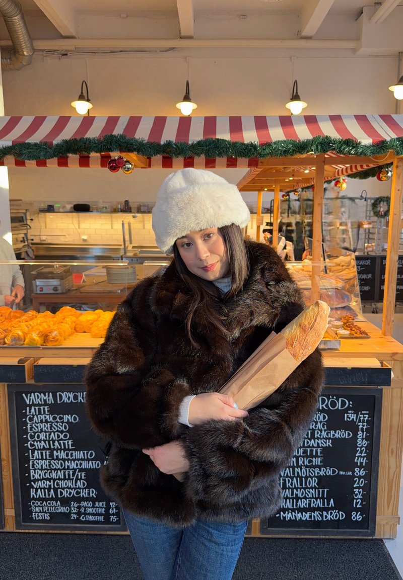 Kobieta w futrzanym płaszczu i białym kapeluszu trzyma bagietkę na stoisku piekarniczym ozdobionym girlandami i pasiastym daszkiem.