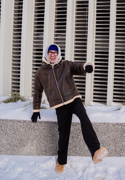 Hombre con chaqueta de invierno, guantes y sombrero equilibrándose en una pierna sobre la nieve cerca de un edificio con persianas verticales en las ventanas.