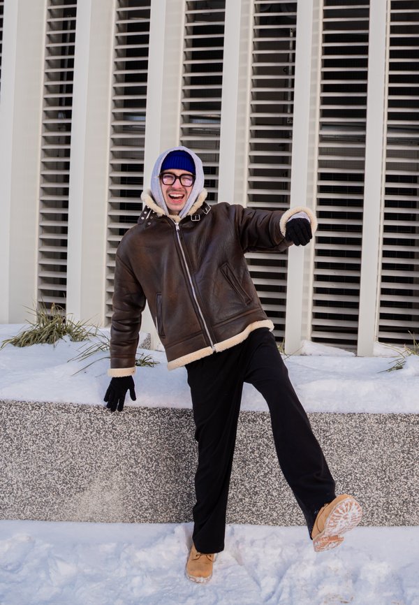 Homme en veste d'hiver, gants et bonnet, en équilibre sur une jambe dans la neige près d'un bâtiment avec des stores verticaux aux fenêtres.