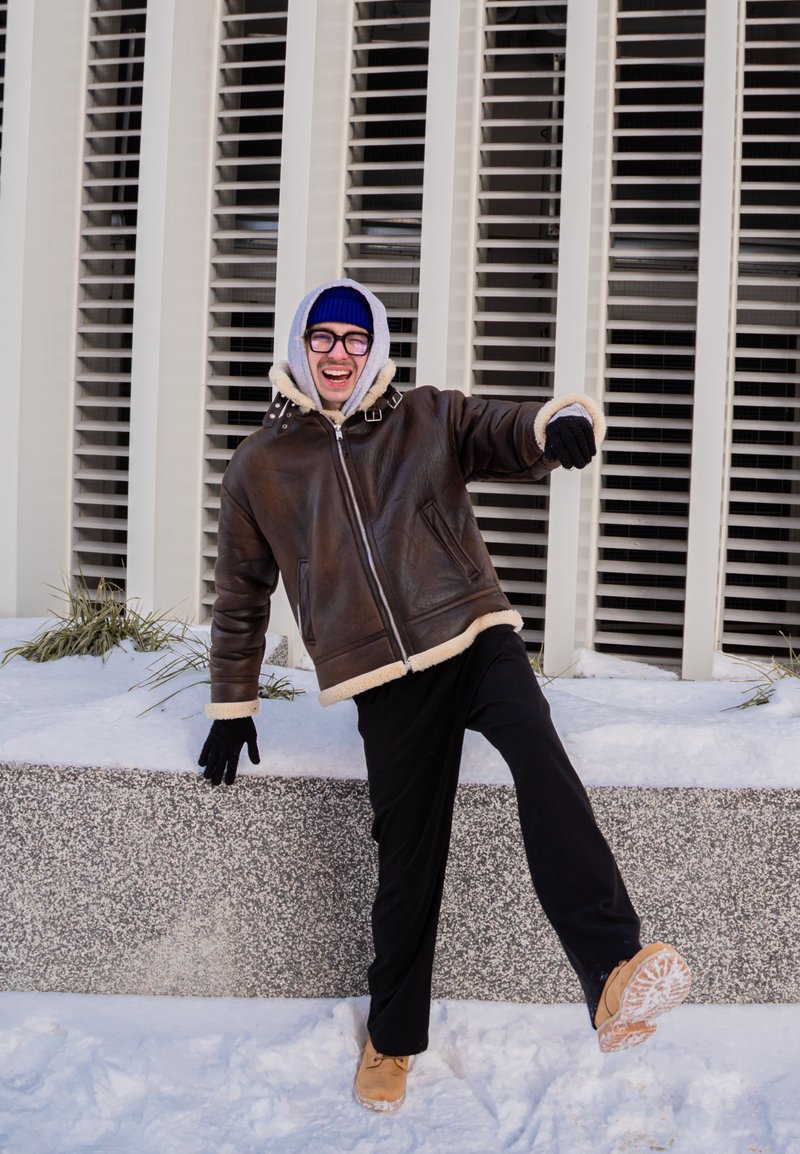 Homme en veste d'hiver, gants et bonnet, en équilibre sur une jambe dans la neige près d'un bâtiment avec des stores verticaux aux fenêtres.