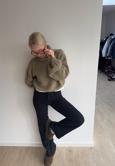 Suéter de punto de gran tamaño en verde oliva, con cuello alto, combinado con pantalones negros ajustados y botas de tacón marrones. Sostiene unas gafas, posando con una pierna levantada.