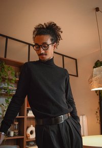 Jeune homme aux cheveux bouclés et lunettes, portant un col roulé noir et une ceinture, debout dans une pièce chaleureuse avec des étagères et des plantes.