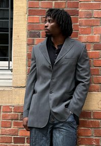 Blazer gris texturizado con dos botones y un bolsillo en el pecho, llevado sobre una camisa negra, combinado con jeans azul oscuro frente a una pared de ladrillo.