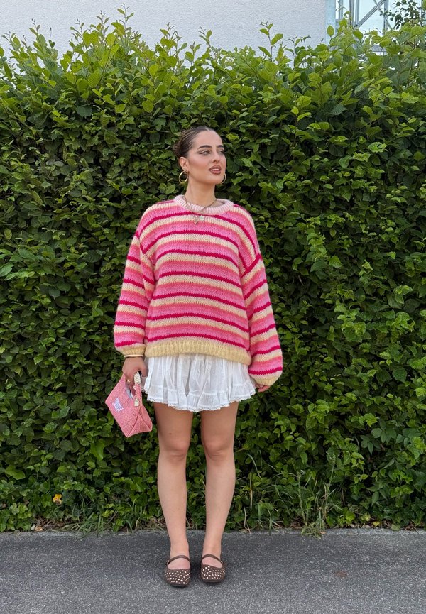 Femme portant un pull rayé rose, une jupe blanche, des chaussures marron, tenant un petit sac rose, debout devant une haie feuillue verte.