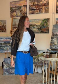 Uma mulher com uma camisa branca e saia azul segura um telemóvel e uma bolsa, ao lado de pinturas coloridas em um estúdio de arte.