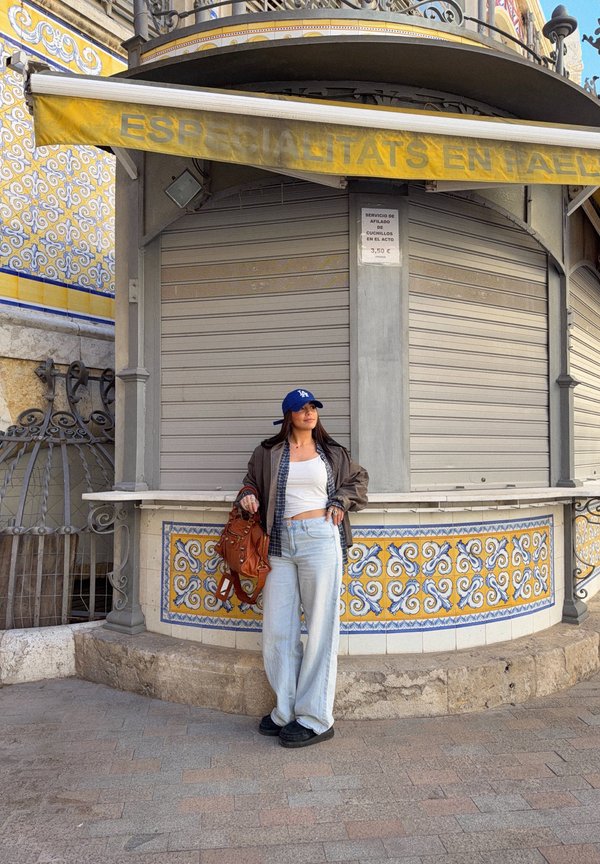 Jonge vrouw met een blauwe pet en casual kleding staat bij een betegeld kiosk in de hoek met gele en blauwe patronen en gesloten luiken.