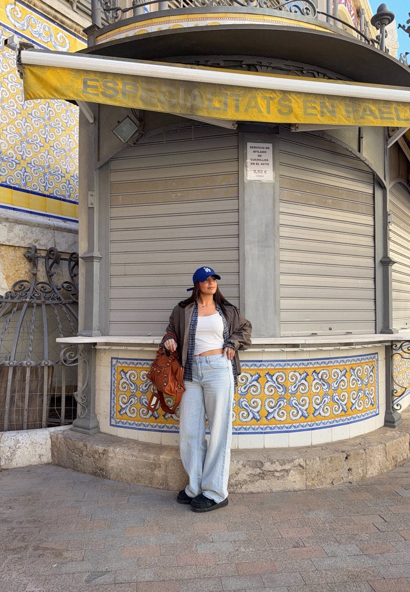 Jeune femme portant une casquette bleue et des vêtements décontractés, posant contre un kiosque d'angle carrelé aux motifs jaunes et bleus avec des volets fermés.