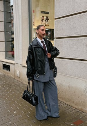 Sort læderjakke over et gråt jakkesæt, hvid skjorte og slips; holder en lille sort håndtaske. Bredbenede bukser og spidse sko.