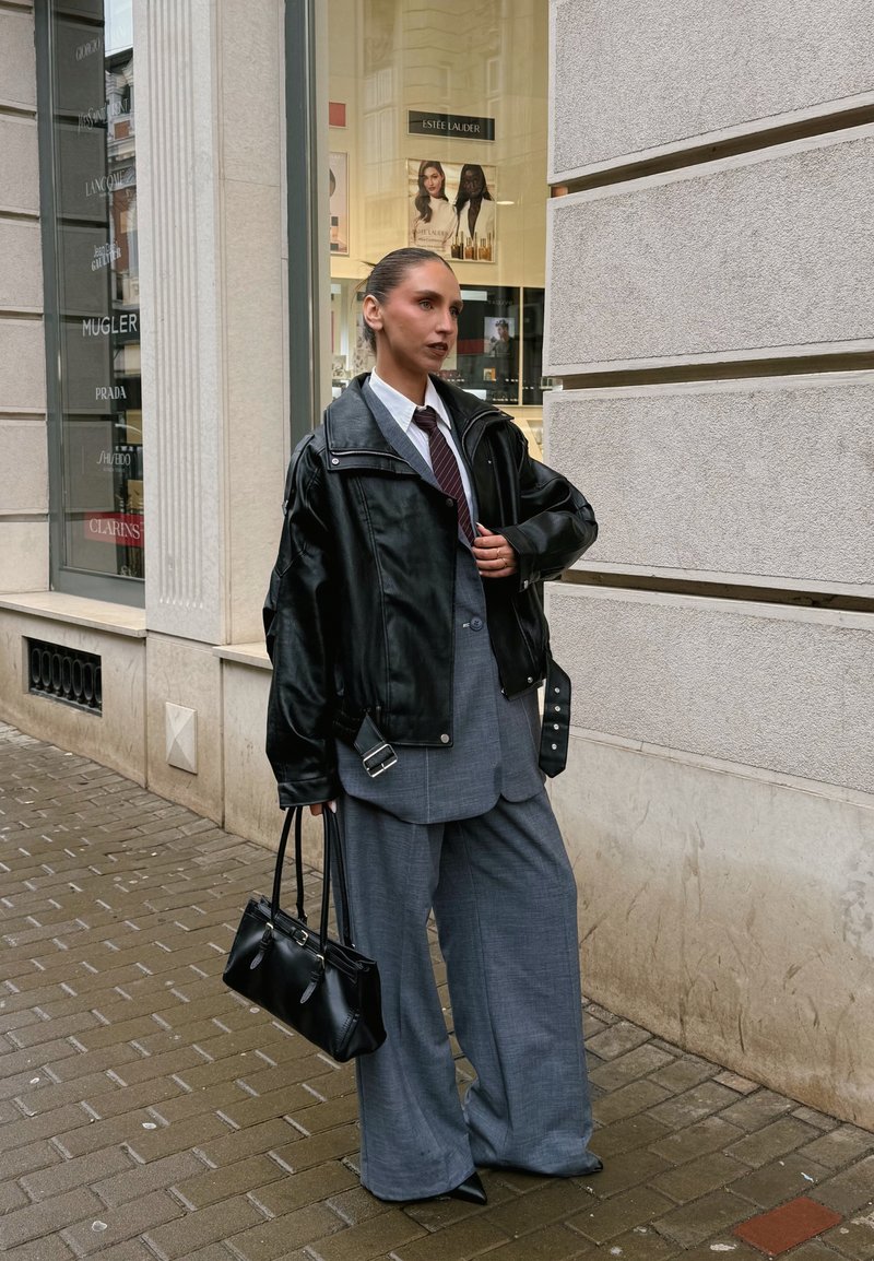Schwarze Lederjacke über einem grauen Anzug, weißem Hemd und Krawatte; hält eine kleine schwarze Handtasche. Weit geschnittene Hosen und spitze Schuhe.