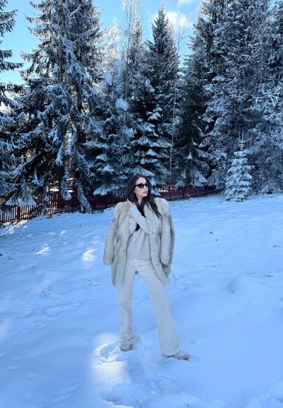 Abrigo de piel sintética en color beige claro, superpuesto a un suéter crema. La modelo lleva pantalones blancos ajustados y gafas de sol negras, con un fondo de bosque nevado.