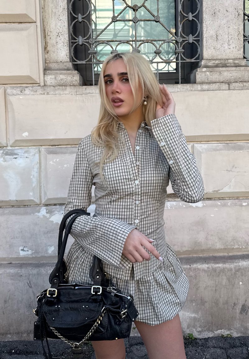 Blondynka w sukience w kratę z zapiętymi guzikami, poprawiająca włosy, trzymająca czarną torebkę, stojąca na tle starego kamiennego muru z ozdobną kratą okienną.