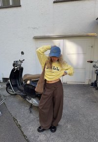 Giovane con felpa gialla, cappellino blu, pantaloni larghi marroni e scarpe nere in piedi accanto a uno scooter nero in un ambiente urbano all'aperto.