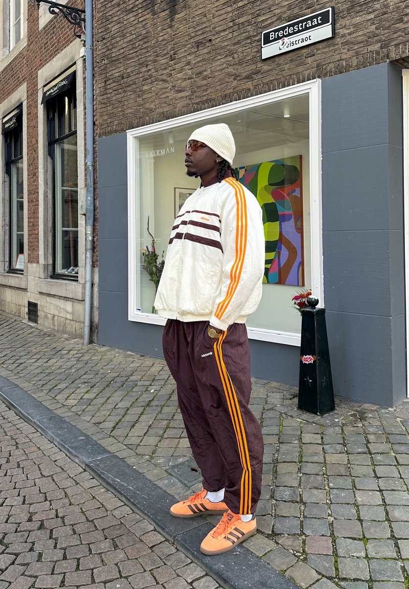 Λευκός και καφέ φούτερ με φερμουάρ με πορτοκαλί ρίγες, σε συνδυασμό με καφέ φόρμες που διαθέτουν πορτοκαλί ρίγες στο πλάι και πορτοκαλί αθλητικά παπούτσια.