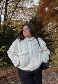 Jachetă albă din sherpa cu accente argintii lucioase, închidere cu nasturi și buzunare laterale; purtată cu pantaloni negri și ochelari de soare într-un decor de toamnă.