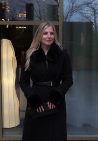 Long manteau noir avec col et poignets en fourrure, ceinturé à la taille. Tient une pochette noire texturée. En arrière-plan, un luminaire.