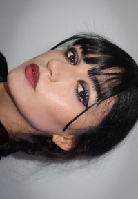 Primo piano di un volto con capelli scuri e frangia, che presenta una pelle evidenziata, labbra rosse e un trucco per gli occhi audace con eyeliner viola e ciglia lunghe.