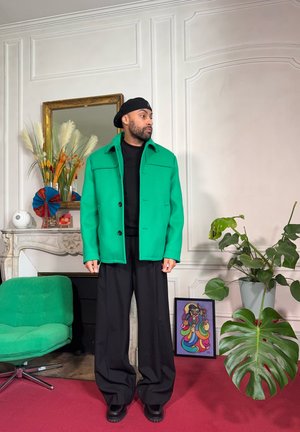 Hombre con chaqueta verde, pantalones negros y gorra negra de pie en una habitación con plantas, arte colorido y una silla verde sobre una alfombra roja.
