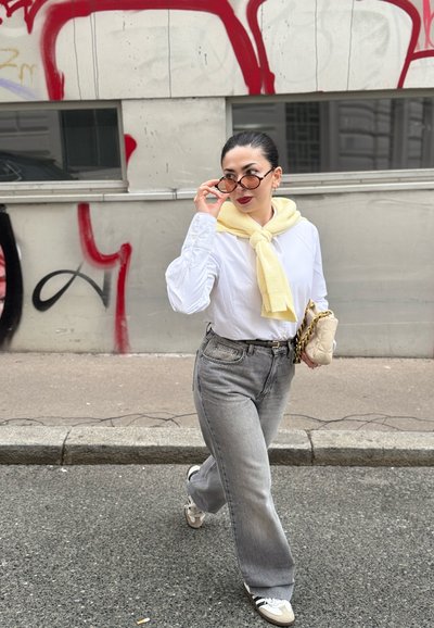 Mujer vestida con una camisa blanca y jeans de pierna ancha grises, con una bufanda amarilla alrededor del cuello. Sostiene un bolso color crema y lleva gafas redondas.